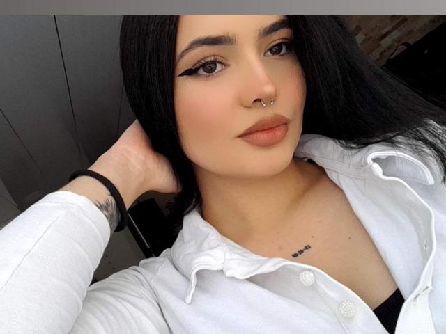 ValeriJoness tem cabelo preto, um piercing no nariz e uma tatuagem no peito. Ela está vestindo uma camisa branca e olhando para a câmera com uma expressão séria..