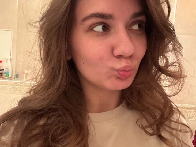EmilyEwexim with pouty lips, brown wavy hair, and a slightly red nose, wearing a white shirt, looking to the side. Her tits are not visible.