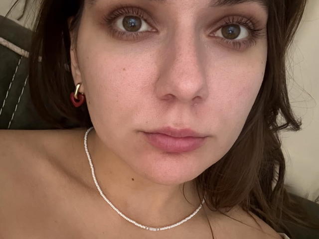 EmilyEwexim's face, showing brown eyes, pinkish lips, small nose, light skin, brown hair, red earrings, and a delicate silver necklace, with bare shoulders visible.