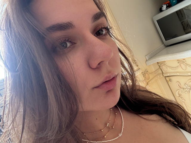 EmilyEwexim with long brown hair, light skin, subtle makeup, and a slight pout, wearing a gold necklace, has small tits, and is looking into the camera.