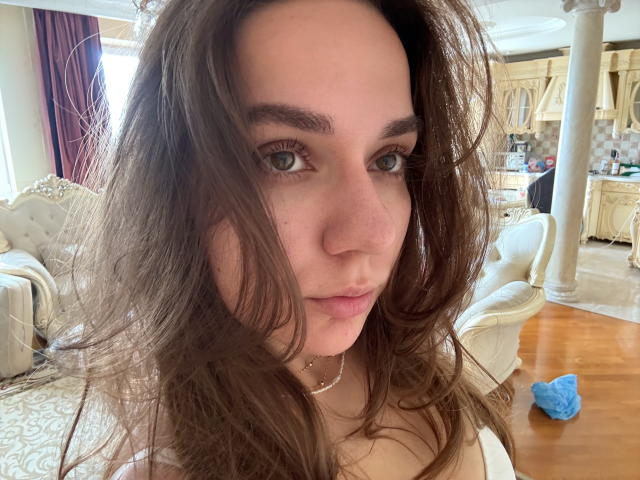 EmilyEwexim has wavy brown hair, green eyes, and a slight pout, wearing a white top, with a blue cloth on the wooden floor in a luxurious room.