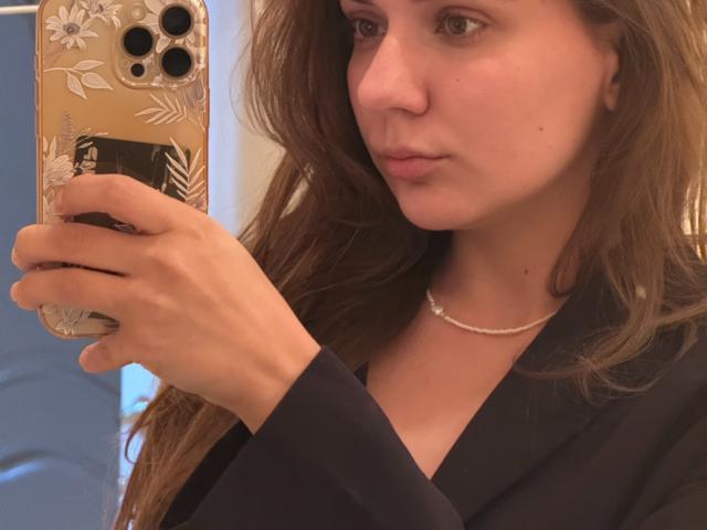 EmilyEwexim with long brown hair, wearing a navy blue shirt and pearl necklace, holding a phone with a floral case, taking a mirror selfie, showing a neutral expression.
