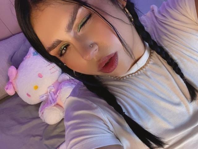 EmmaMyles with one eye closed, black braids, green eye makeup, nose piercing, pink lips, white ribbed shirt, holding pink Hello Kitty stuffed toy, looking seductive.