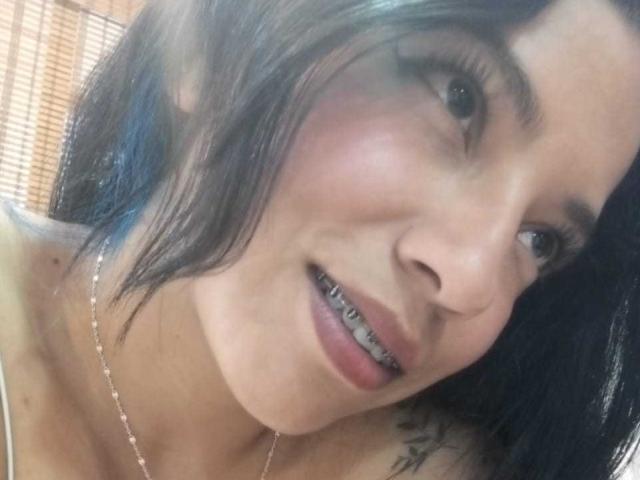 PaulinaColemann with black hair, fair skin, and a silver necklace, showing metal teeth in a slightly open-mouthed smile. Her tattoo is partially visible on her right shoulder.