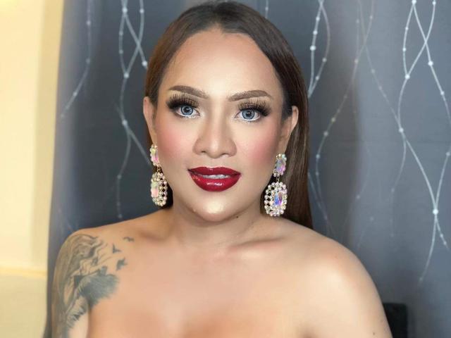 MissAthene has blue eyes, red lipstick, and a tattoo on her left shoulder. She's wearing diamond earrings and has long brown hair. Her boobs are partially visible.