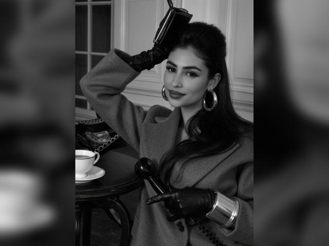 Fotografia in bianco e nero di MiilaWx con capelli scuri, che indossa un cappotto, guanti di pelle e orecchini a cerchio, mentre tiene un telefono, con una tazza su un tavolo in primo piano..