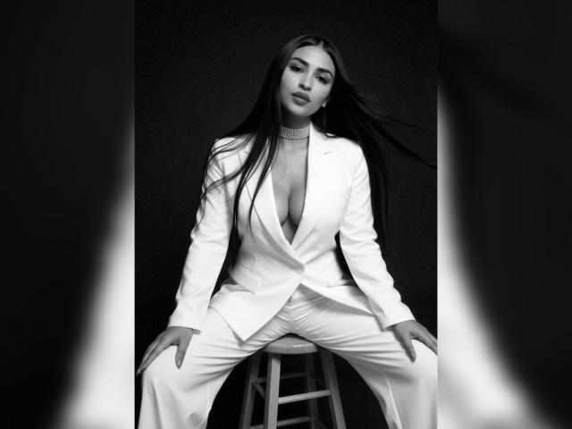 MiilaWx, sitting on a stool, wearing a white blazer and pants, with a deep V-neck exposing her large tits, long dark hair, and a confident, sexy expression. Black-and-white photo.