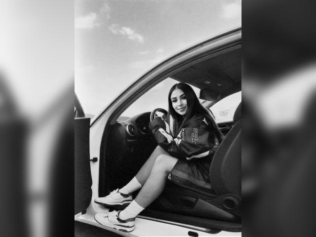 MiilaWx with long dark hair, sitting in a car, wearing a jacket, shorts, socks, and Nike sneakers, looking down, hands on knees, car door open, black-and-white photo.