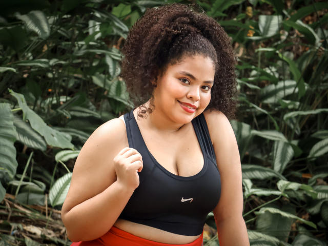 TaniaBelle has curly brown hair, tan skin, and red lipstick. She's wearing a black sports bra with visible tits, orange pants, and smiling with a hand near her chest.