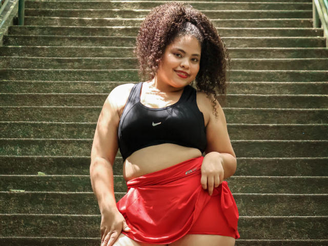TaniaBelle has curly brown hair, a black Nike sports bra, and a red skirt, pulling it up to expose her tan, curvy, bare-ass. She's smiling with a sexy look.