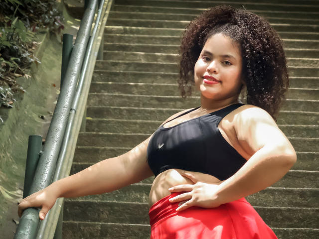 TaniaBelle has curly brown hair, a black sports bra showing her small tits, and a red skirt, standing on a staircase with one hand on a green rail.