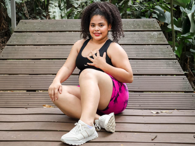 TaniaBelle with curly dark hair, wearing a black sports bra and pink shorts, sitting on a wooden deck, touching her chest, white sneakers, smiling, chubby, curly afro.