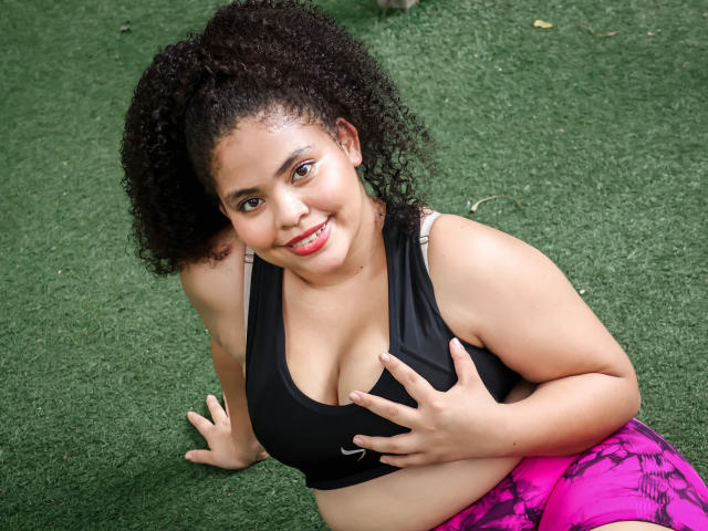 TaniaBelle, a curvy woman with curly black hair, smiling at the camera, wearing a black sports bra and pink patterned pants, touching her big boobs on green grass.