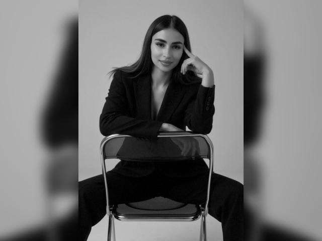 Schwarz-weißes Foto von MiilaWx mit langen, dunklen Haaren, die auf einem Metallstuhl sitzt, einen schwarzen Blazer und eine Hose trägt, ihre Hand auf ihrem Kinn ruht und lächelt..