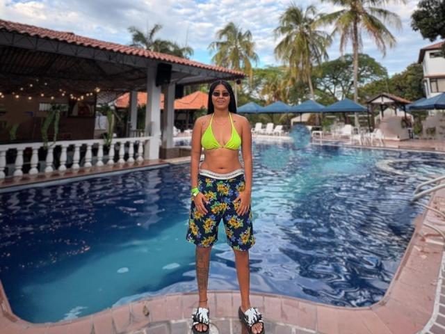 KathiaLewis, tan-skinned, with small tits, in a neon yellow bikini top and colorful floral shorts, stands by a blue pool, wearing black and white sneakers, smiling.