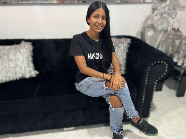 KathiaLewis with long black hair, smiling, wearing a black Mischi t-shirt, ripped blue jeans, and black sneakers, sitting on a black velvet couch with studded edges.