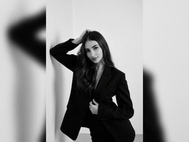 MiilaWx with long dark hair, wearing a black suit, standing against a wall, hand in hair, hand on chest, black-and-white photo, soft smile, sexy, confident.