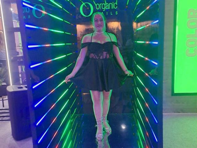 TsKami stands in front of a neon-lit wall, wearing a black off-shoulder dress with cutouts, showing her tits, and lace-up heels, smiling with arms slightly spread.