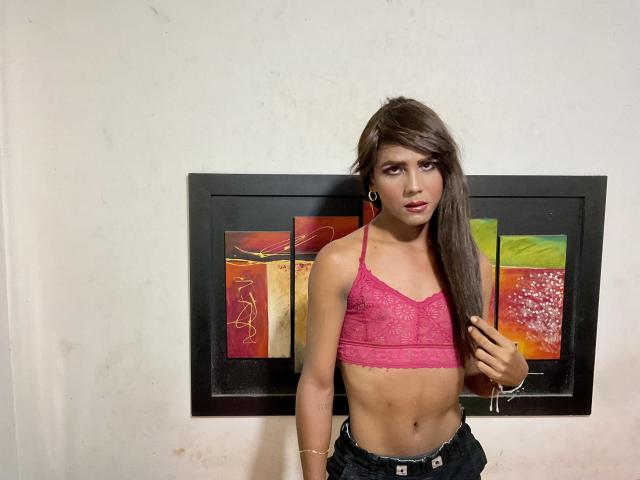 MarilynbrownX with long brown hair, pink lace bra, small tits, slim, wearing dark jeans, standing in front of abstract wall art, holding hair with right hand.