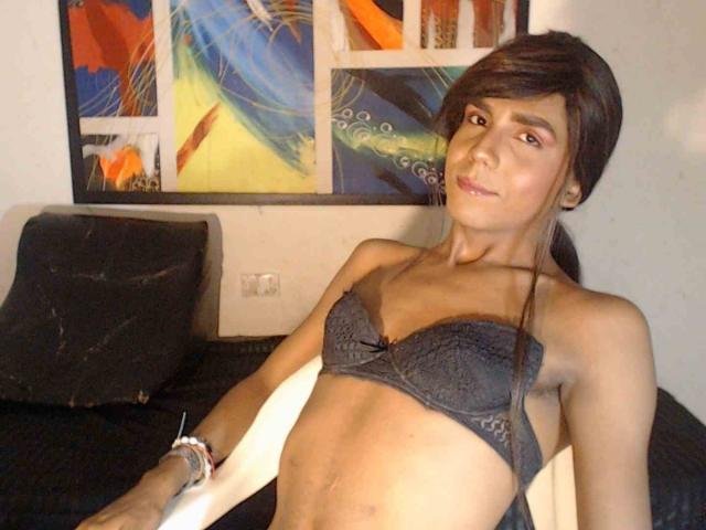 MarilynbrownX with brown hair, wearing a grey lace bra, has small tits and a slim, toned body, sitting on a black couch, looking at the camera with a slight smile.