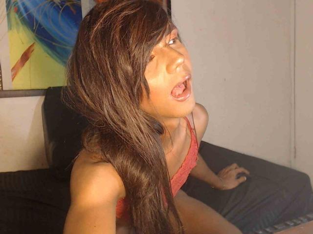 MarilynbrownX with long brown hair, open mouth, red tank top, sitting on black bed, looking up with tongue out, warm light on face, right hand on bed.