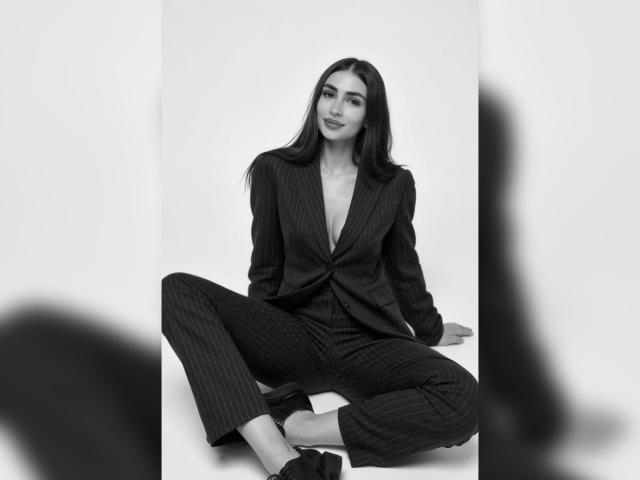MiilaWx, con lunghi capelli scuri, indossa un completo a righe e siede con le gambe divaricate, rivelando un profondo scollo a V, e porta dei tacchi alti neri, in una fotografia in bianco e nero..