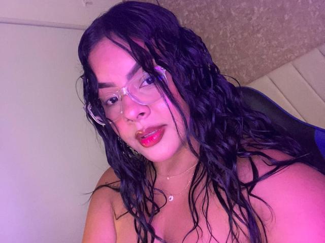 AmarantaCarey with wet black hair, wearing clear glasses and pink lipstick, has red lips and a small heart necklace, showing her bare shoulders and a cute face.