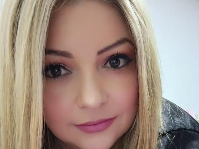 NatalyHami has blonde hair, blue eyes with dark eyeliner, pink eyeshadow, and pink lipstick. She's smiling with a slight blush on her fair skin. Her nose is small.