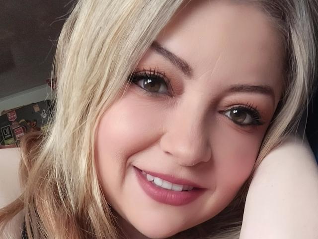 NatalyHami's face, smiling with pink lipstick, green eyes, and blonde hair, head resting on arm, showing cute, sexy, and attractive features. She has a small nose and visible eyelashes.