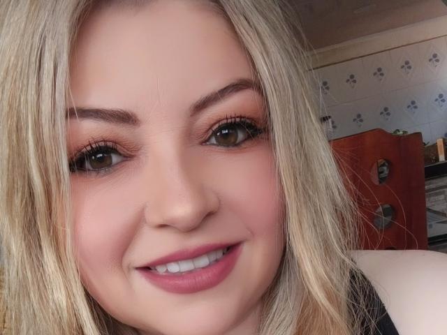 NatalyHami has blonde hair, brown eyes, and pink lipstick. She's smiling with her tits slightly visible. Her makeup includes orange eyeshadow and long eyelashes.