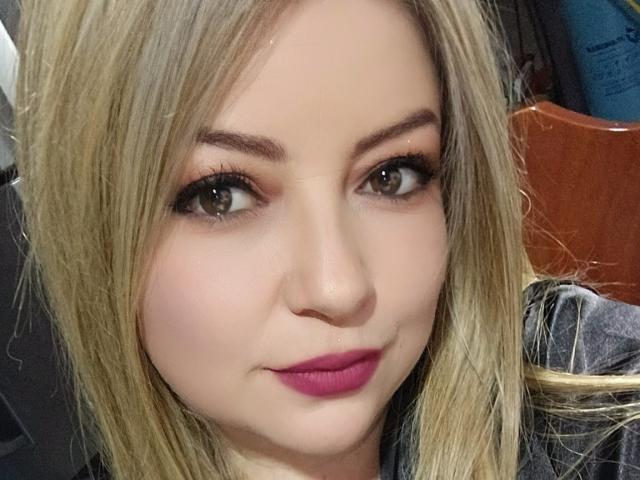 NatalyHami has blonde hair, brown eyes, pink lipstick, and a slight smile. Her makeup includes long eyelashes and subtle eyeshadow. She has a cute, sexy face.