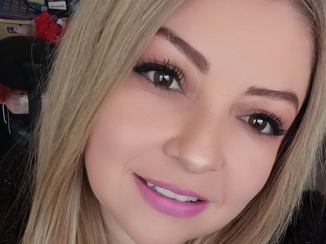 NatalyHami has blonde hair, brown eyes, dark eyeliner, thick eyebrows, pink lipstick, and a slight smile. Her tits aren't visible, just her face.