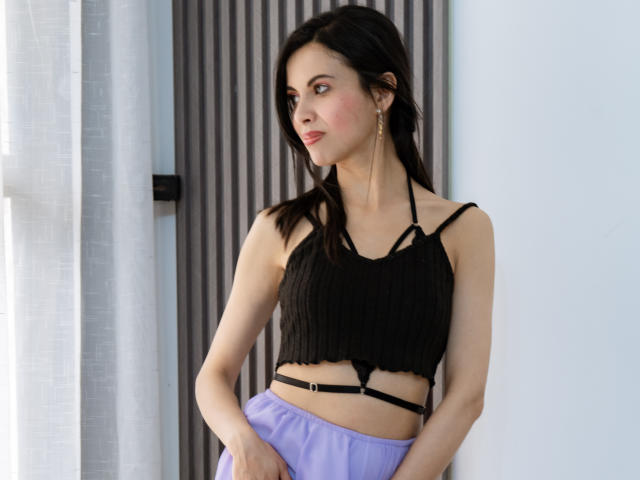 TaylaCooperk with black ribbed crop top, showing small tits, black strappy details, and purple pants, standing near white curtain, looking sideways, wearing gold earrings.