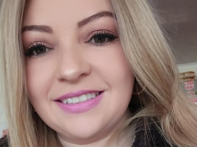 NatalyHami's face with blonde hair, pink lipstick, and a smile, showing her white teeth, black eyeliner, and brown eyes. She's wearing a black top.