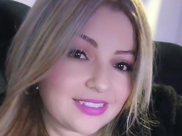 NatalyHami has blonde hair, pink eyeshadow, and purple lipstick, smiling with white teeth, and has a small nose ring, showing cute, sexy, and attractive features.