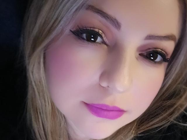 NatalyHami's face with blonde hair, pink blush, black eyeliner, purple lipstick, and dark eyes. She's looking at the camera with a slight smile. Her nose is small and cute.