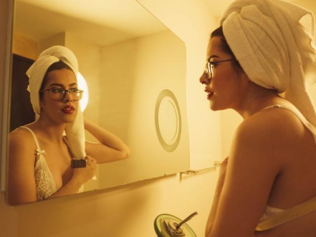 BurningPleasures with a towel on her head, glasses, and white bra, looking at herself in a mirror, showing her tits and ass in a warm, yellow-lit bathroom.