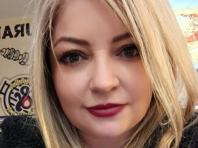 NatalyHami has blonde hair, green eyes, and red lipstick, with a slight smile and a black fuzzy top, looking directly at the camera. Her face is centered.