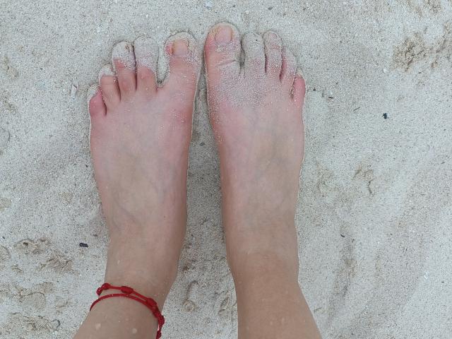 KayleeM's bare feet, slightly dirty with sand, standing on white beach sand, pinkish toes, red string bracelet on left ankle, visible veins, slight redness on toes.
