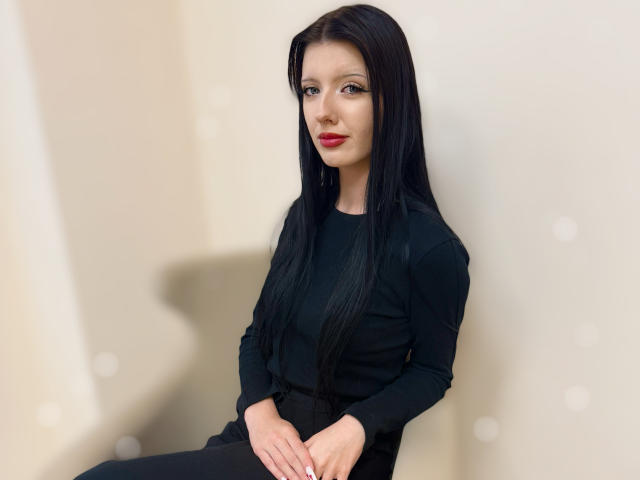 RoxyHopper met lang zwart haar, gekleed in een zwarte top met lange mouwen, zittend, handen op zwarte broek, rode lipstick, kijkt naar de camera, bleke huid, subtiele make-up..
