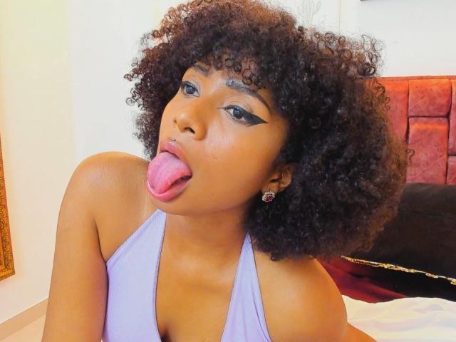 MeganWilliams with curly black hair, purple halter top, and nose ring, sticking out her tongue, showing black winged eyeliner and pink tongue. Her small tits are visible.
