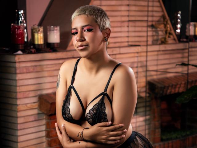 DulceSweett met kort blond haar, roze oogschaduw, een zwarte kanten bh, waarbij haar borsten zichtbaar zijn, armen gekruist, een tatoeage op haar linkerarm, oorbellen, staand in een zwak verlichte kamer, met een glimlach..