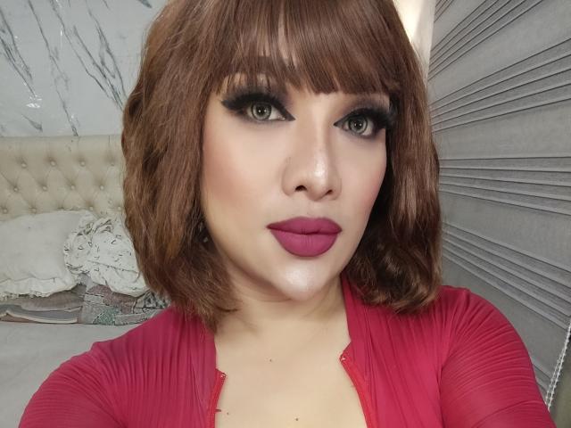 MissAthene with shoulder-length brown hair, heavy black eyeliner, and magenta lipstick, wearing a red top, showing off her puffy tits, in a selfie.