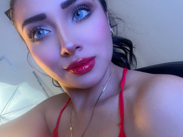 LillyTaylor has shiny red lips, blue eyes, and a slight smile. She's wearing a red lace top and a gold necklace. Her dark hair is pulled back.