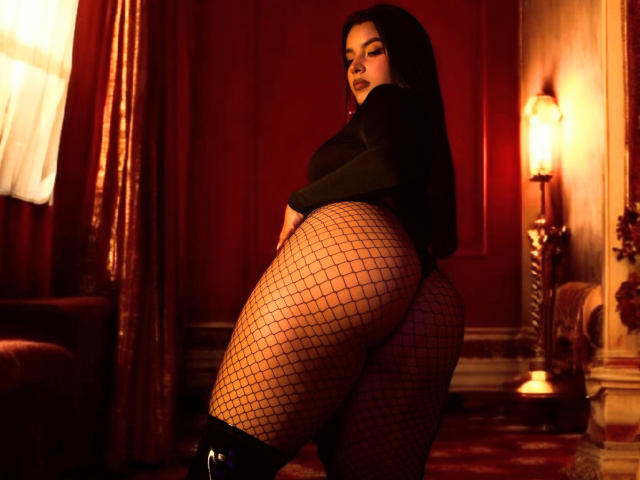 ArtemisaStone with dark long hair, black top, and fishnet stockings, showing off her big ass, standing in a dimly lit room with red walls and a lamp.