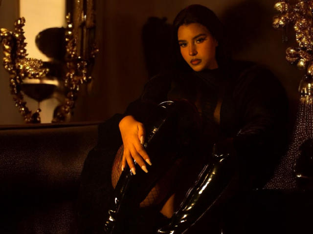 ArtemisaStone with dark hair, black sheer top, and shiny black boots, sitting on a dark leather couch, hands on her knees, looking sultry with a serious expression.