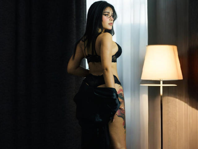ArtemisaStone with black lace bra and panties, holding a black jacket, showing a colorful tattoo on her right thigh, standing next to a lit lamp, looking back.
