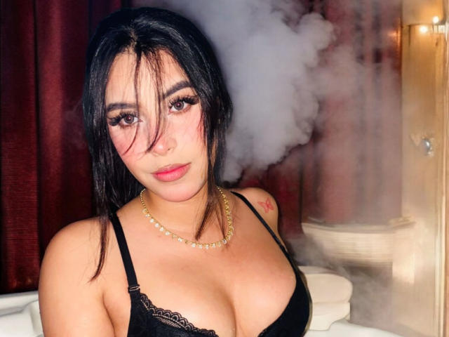 ArtemisaStone with black lace bra, gold necklace, and pink makeup, has her tits on display, with steam in the background and her nose ring visible.