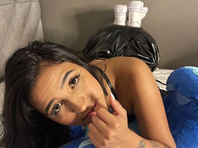 VixenScarlet mit dunklem Haar, braunen Augen und gebräunter Haut, liegt auf einem Bett, lutscht an ihrem Finger und trägt einen schwarzen, glänzenden Latex-Hosenanzug, ein blaues Stofftier und weiße Socken..