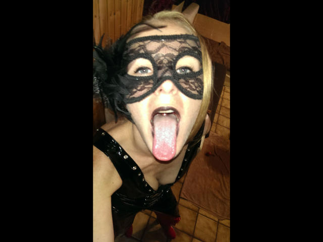 HeisseLina-hot with blonde hair, black lace mask, tongue out, and black sequined top, showing her pink tongue and slightly open mouth in a wooden room.